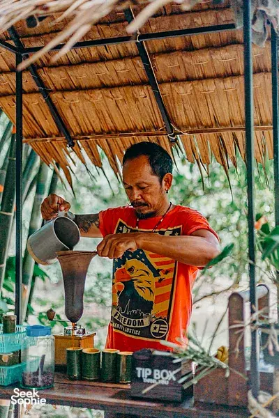 Community stall worker pouring from a large kettle under a palm roof in Khao Lak for a conservation-focused tour
