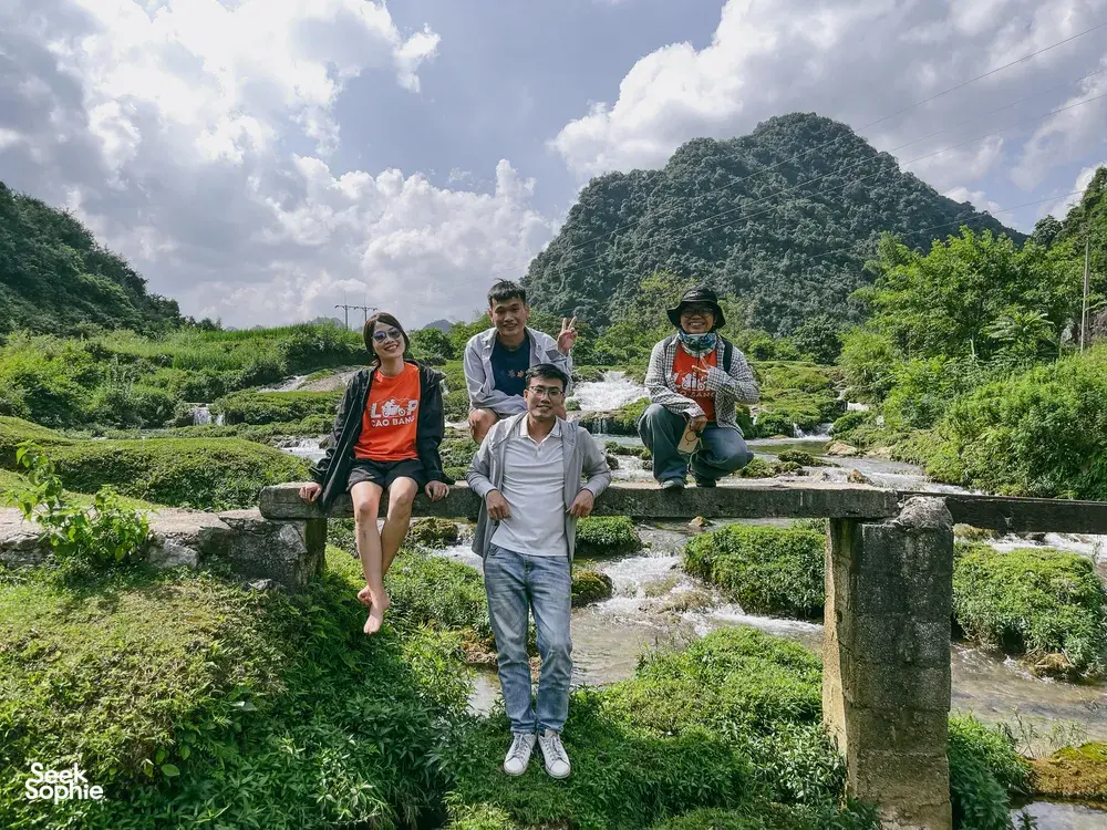 Helmets on, a group of motorbike riders posing on grassy stone steps beside a stream in Cao Bang, Vietnam, on the loop