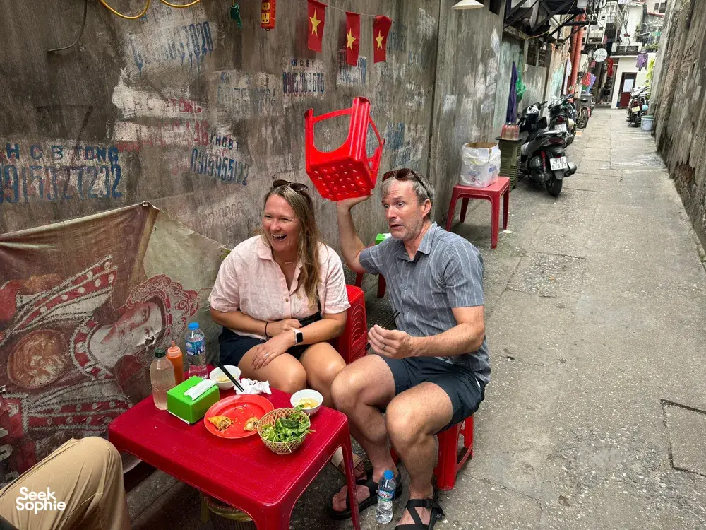 Travellers seated at a low red table eating in a narrow Hanoi laneway, with dishes, drinks and street clutter