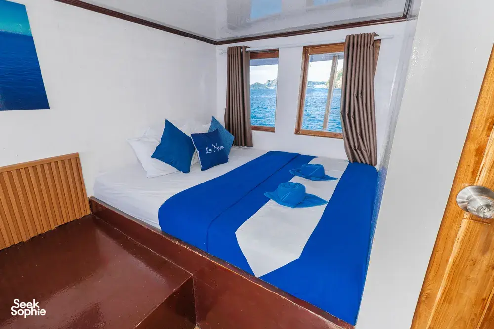 La Famille cabin with neat blue bedding, window curtains and sea water visible beyond, Komodo boat trip, Labuan Bajo