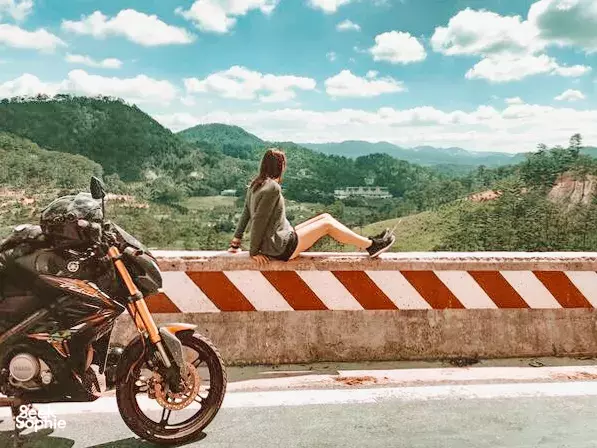 Woman sitting on a striped roadside barrier beside a parked motorbike, gazing over green valleys on the Easy Rider route