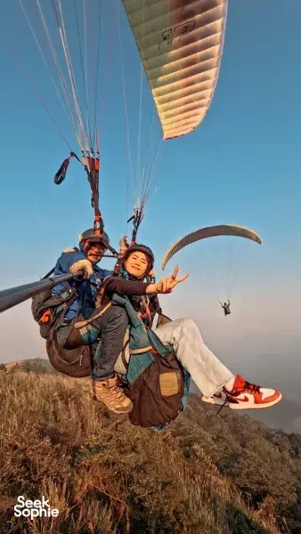 Passenger waving from a tandem paraglider harness as the canopy fills above on a launch hillside near Hanoi, holding steady