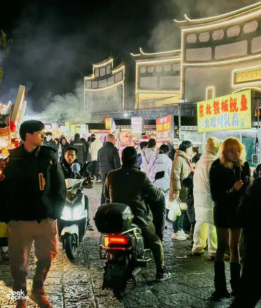 Bustling Lijiang night market crowd walks past neon-lit stalls and scooters, setting the scene for a local food hunt