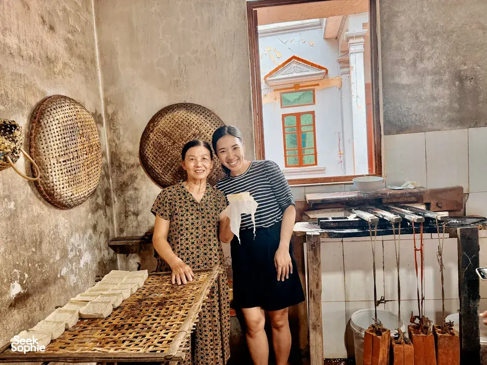 Two women smiling in a Hanoi tofu-making kitchen beside bamboo mats of pressed tofu, with doorway light behind them