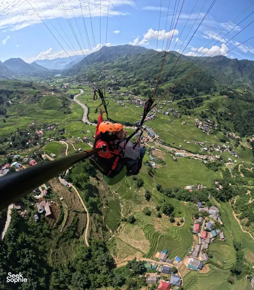 View from a paraglider seat as the canopy lines stretch over a valley with a winding river and village clusters in Sapa