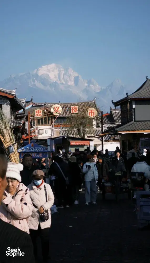 Crowded narrow market street in Lijiang old town with traditional shops, Chinese signboards, and snow-dusted mountains in view