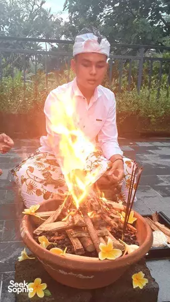 ADDA Yoga host tending a Balinese fire ritual beside a flower-strewn clay brazier in a garden courtyard in Canggu