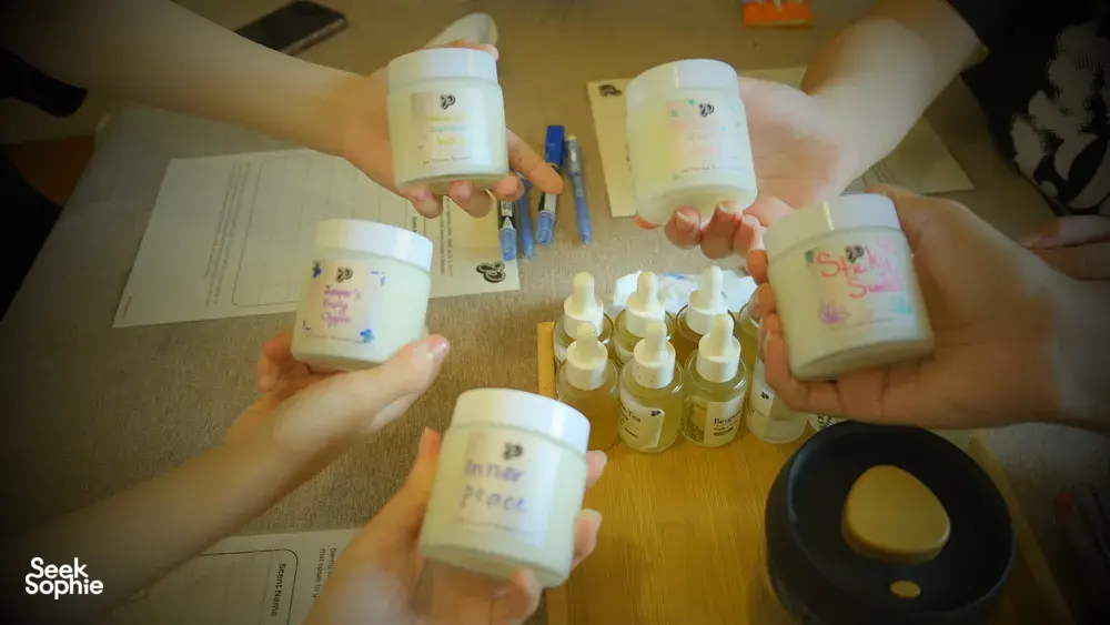 Hands raised holding labelled coco-soy candle jars over a table of oils and wicks during a Singapore candle workshop