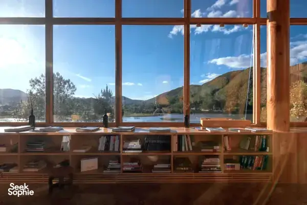 Sunlit reading area inside a wooden retreat room, with books on shelves and hills visible through floor-to-ceiling windows