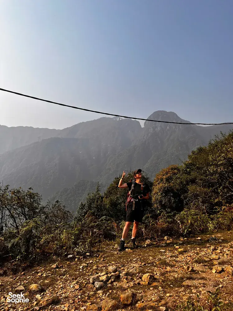 Hiker stands on a rocky ridge trail in Sapa, raising a hand while trekking through sunlit mountain mist