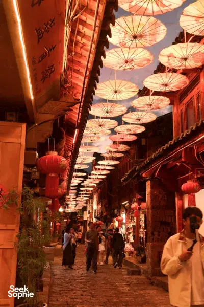 Lantern-covered alley in Lijiang Old Town, warm red light beneath hanging parasols as visitors stroll through the narrow lane