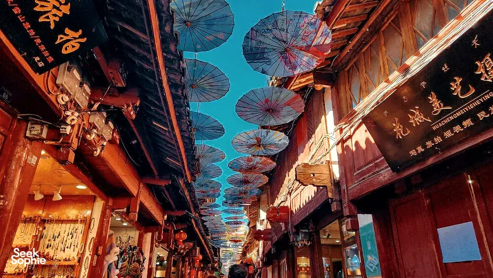 Narrow wooden alley in Lijiang Old Town strung with colourful paper parasols overhead, with shop signs lining both sides