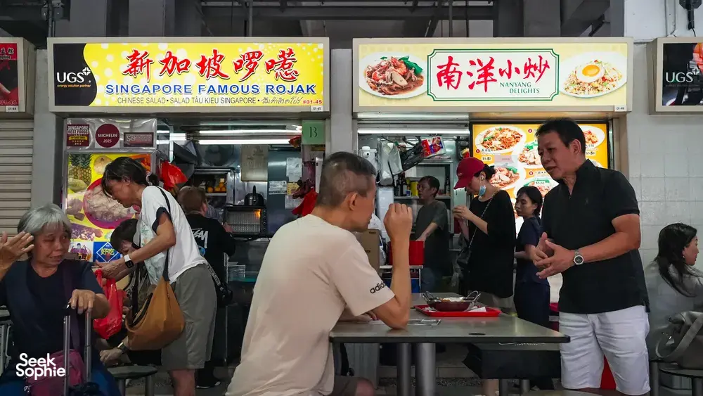 Hawker centre rojak stall in Singapore with cooks preparing food behind the counter as diners wait under menu boards — Singapore hidden gems