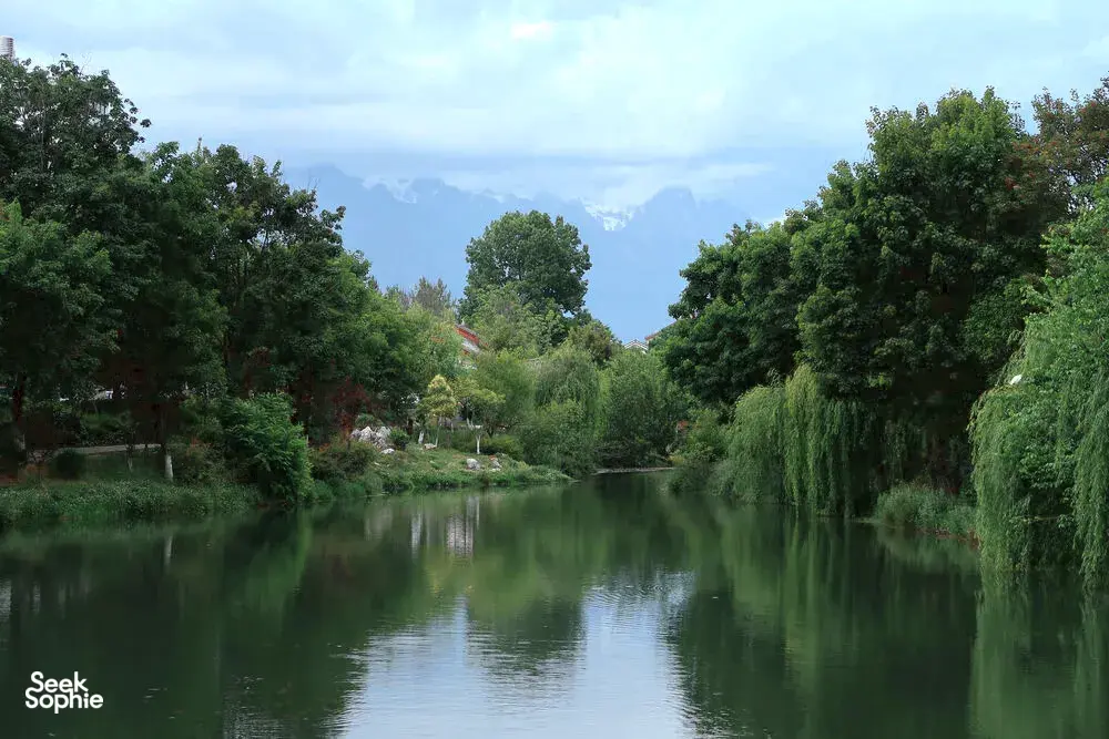 Calm green river in Lijiang framed by weeping trees and garden paths, with distant mountains under a pale blue sky