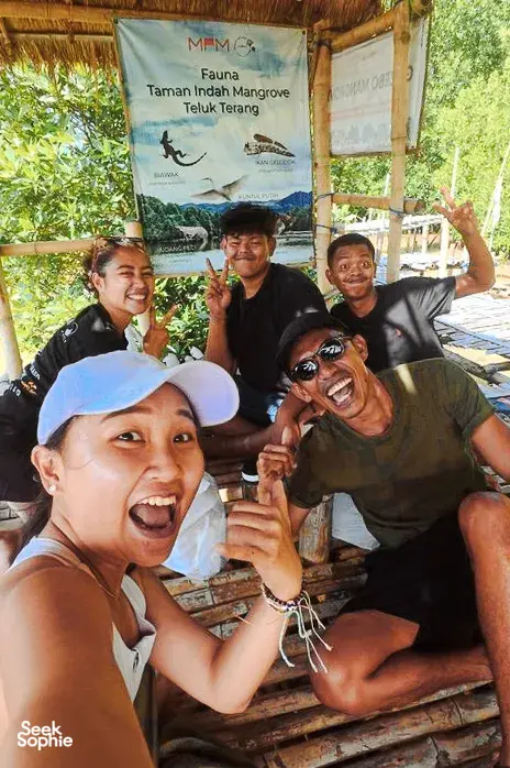 Smiling visitors posing at a mangrove village jetty with a banner for Taman Indah Mangrove during a day trip in Labuan Bajo