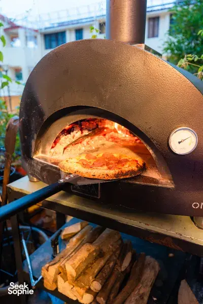 Close-up of a wood-fired pizza turning in an outdoor oven at Secret Pizza Club in Singapore, with firewood below