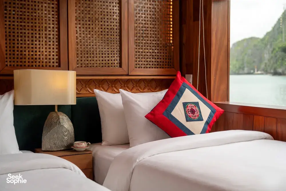 Boutique cruise cabin in Cat Ba with two neatly made beds, reading lamps and pillows, looking out on calm bay waters, Halong Bay