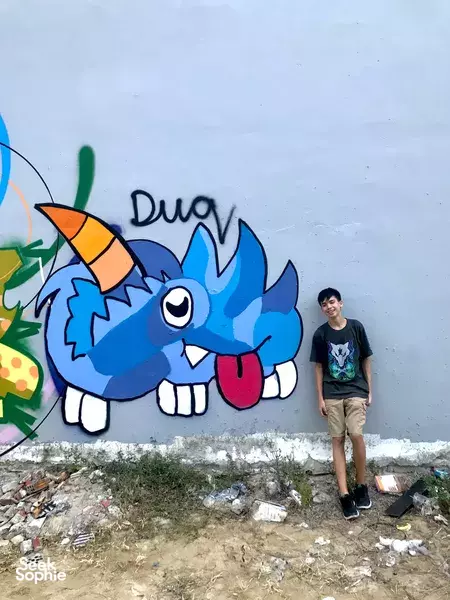 Two people posing by a large blue dragon graffiti wall, one standing nearby and one painting for a creative workshop in Da Nang