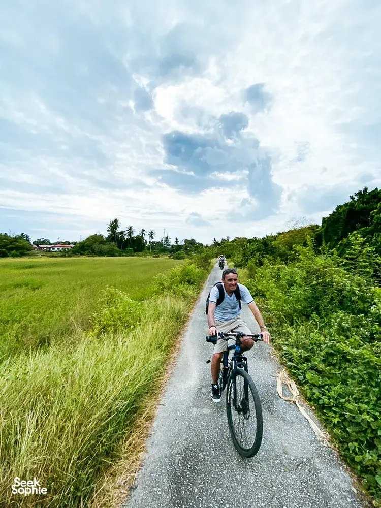 A cyclist rides along a gravel path in Langkawi, through green fields and dense shrubs under a bright sky