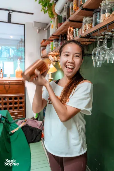 Smiling woman shakes or pours a drink at a Singapore cocktail making class in a cosy nature hideout, with bottles on shelves