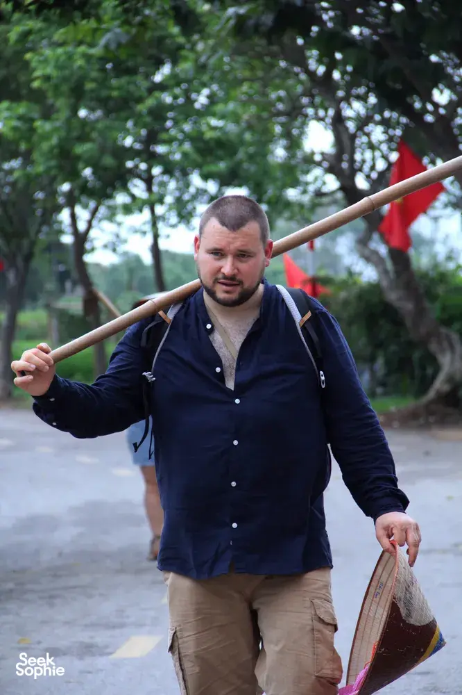 Doai Tour & Homestay host carrying a basket and hoe outdoors along a rural path during hands-on rice farming near Hanoi