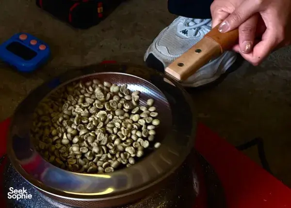 Hand turning coffee beans in a metal pan on a workshop stove, showing the processing stage for Yunnan coffee in Lijiang