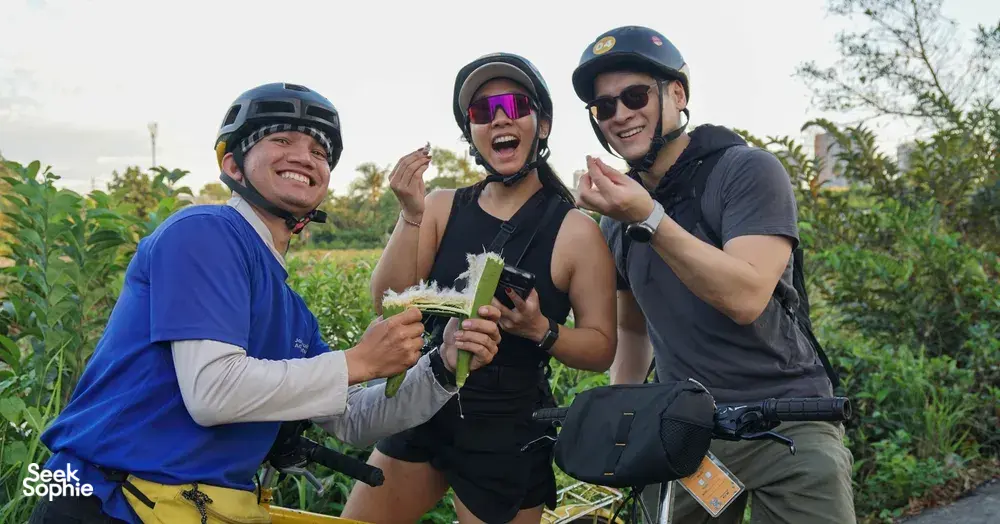 Jackfruit Adventure cyclists in helmets laughing and holding drinks on a bike ride through green paths near Ho Chi Minh City
