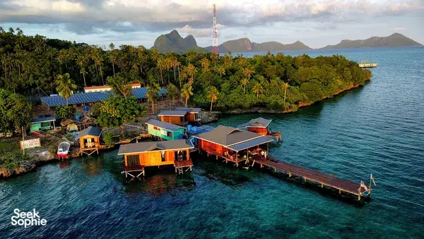 Aerial view of Semporna sea lodge cabins along the shoreline with a long jetty, boats and distant hills under evening light