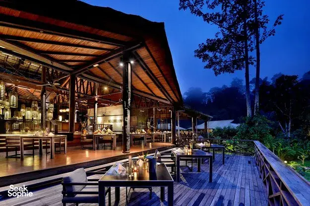 Borneo Rainforest Lodge veranda with dining tables overlooking Danum Valley rainforest at dusk, framed by tall trees and soft lights