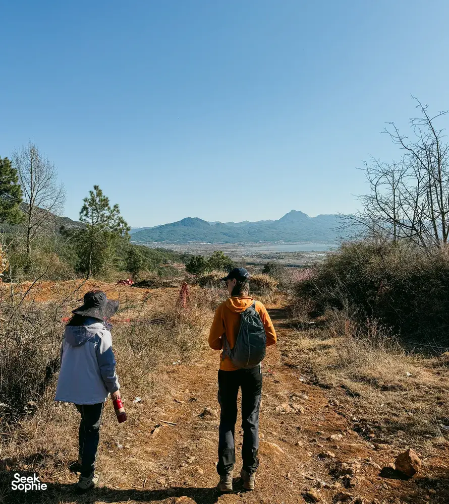 Backpackers trekking along a rural path outside Lijiang, with hazy mountains and a broad valley stretching in the distance