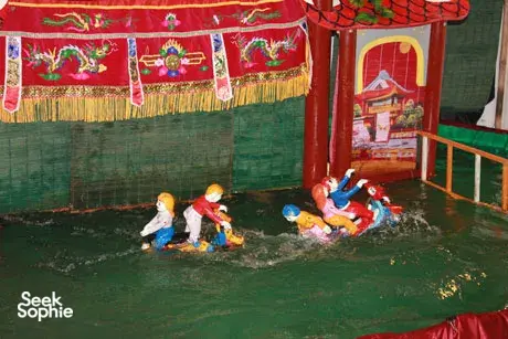 Boneka air kayu berwarna-warni mengapung di panggung tergenang di bawah cahaya lampu dalam pertunjukan tradisional Hanoi