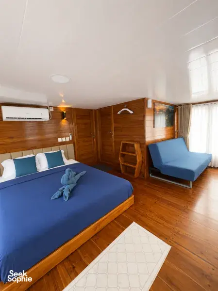Twin beds and a lounge chair in a wood-lined cabin, with sunlight over sea views on a 3D2N shared Komodo cruise, Komodo Island