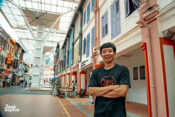 Smiling Triad Trails guide in a narrow Singapore arcade lined with shopfront doors, sharing stories from the ex-gangster era