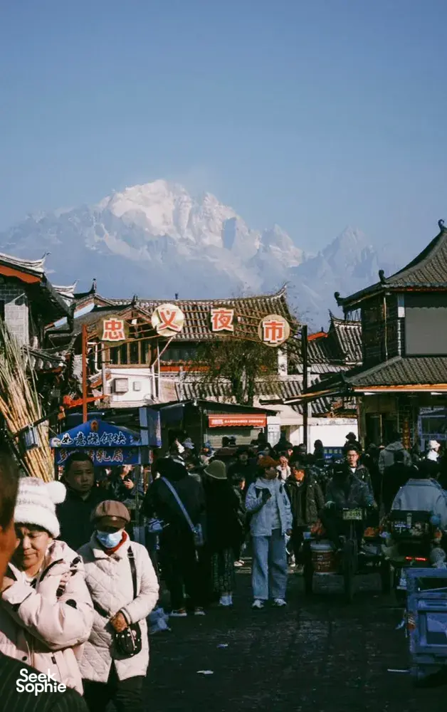 Evening street market in Lijiang with lantern-lit shopfronts, shoppers bundled up, and a snow-capped mountain towering behind