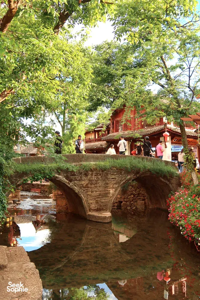 Arched stone bridge over a calm canal with visitors and traditional wooden buildings in Shuhe Ancient Town, Lijiang