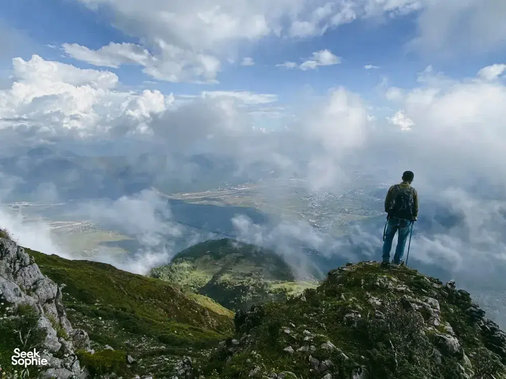 Hiker with trekking poles standing on a rocky ridge as mist and clouds drift over valleys during the Jade Dragon hike