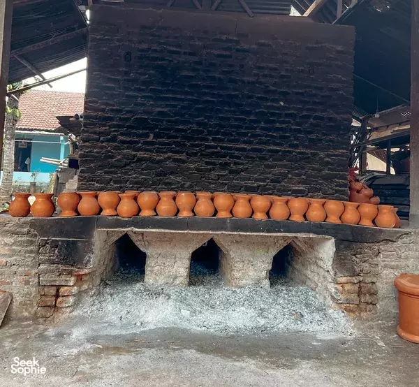 Terracotta pots lined along a brick kiln wall in a Yogyakarta pottery village, with an open firing hearth below