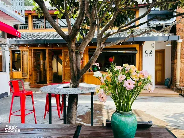 Courtyard dining setup in Lijiang with a stone table, bright red stools, vase of pink flowers and a large banyan tree