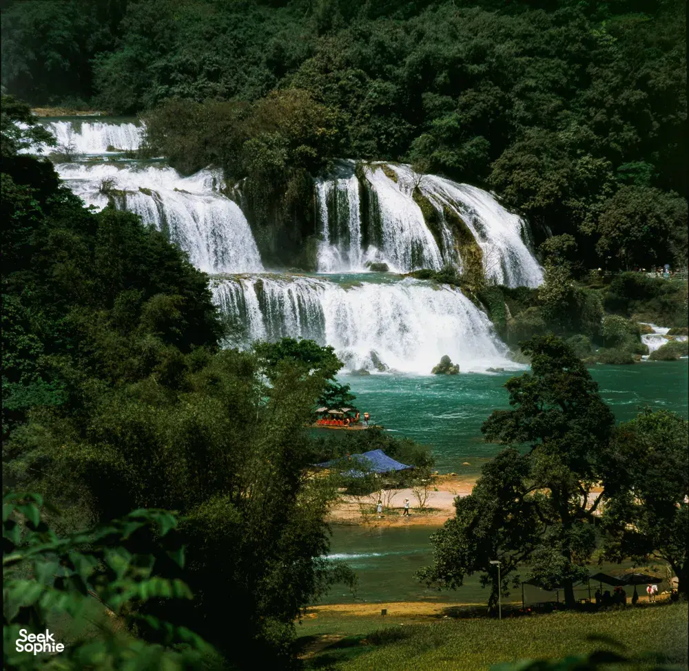 Wide cascade waterfall in a lush green valley in Cao Bang, with boats and visitors along the riverbanks