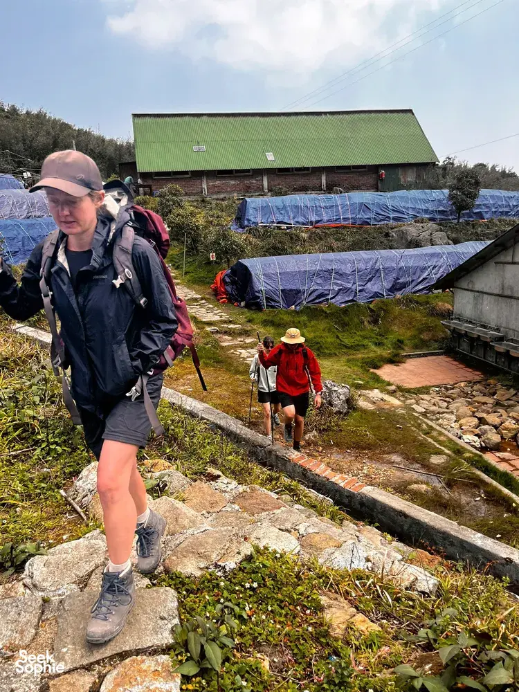 Hiker walking along a stone path past blue tarps and a green-roof shelter in Sapa on the Fansipan eco trek