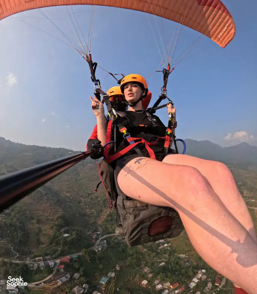 Tandem paragliding close-up of an orange-helmeted passenger reclining in the harness as the pilot glides over valley farmland in Sapa