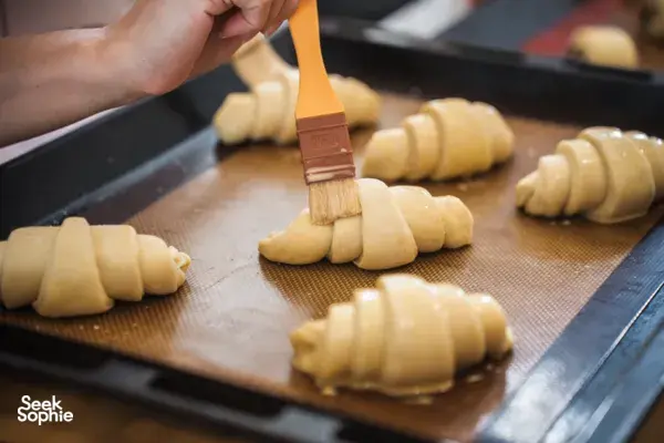Chef brushing egg wash onto rolled croissant dough on a mesh-lined tray at French Cooking Lab’s Singapore class