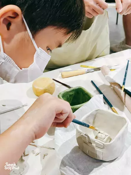 Masked child carving shapes in clay at a kids pottery table in Singapore, working with sculpting tools — workshops in Singapore