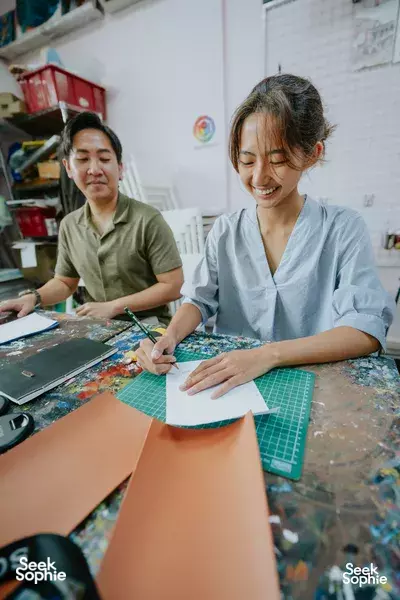 Two people in smocks mark and trim leather journal pages on a cutting mat at Avant-Garde Art Space, during workshops in Singapore