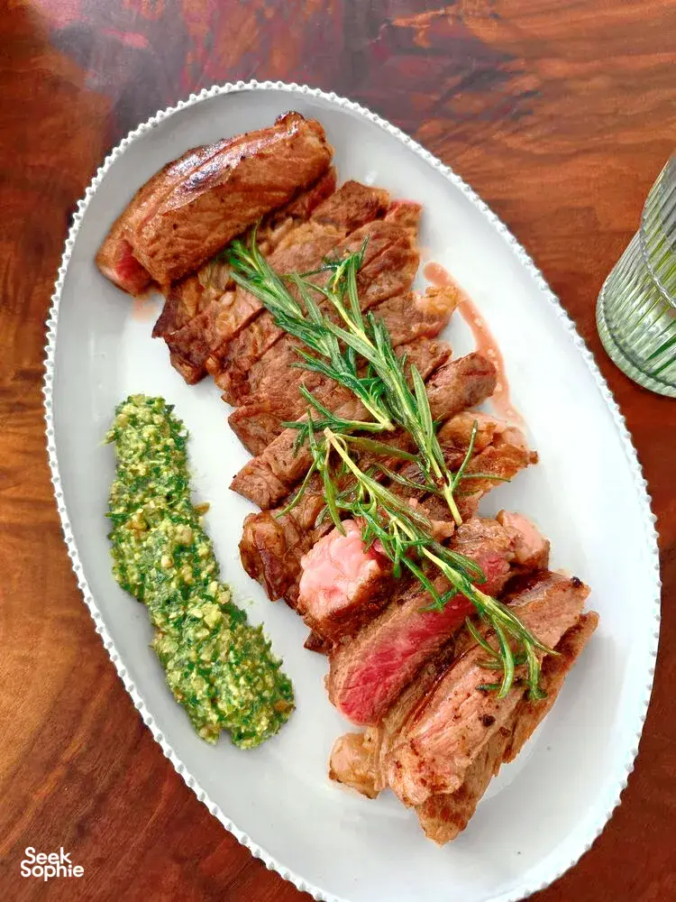 Hirisan daging dengan rosemary di atas pinggan putih bergaya antik, dihidangkan bersama sos herba hijau di sisi