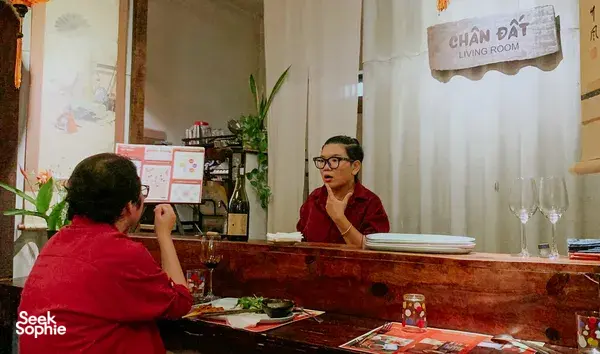Orang ramai sedang menjamu selera di ruang dalaman yang nyaman di Chan Dat Living Room, dengan tuan rumah duduk di belakang kaunter hidangan di Hoi An
