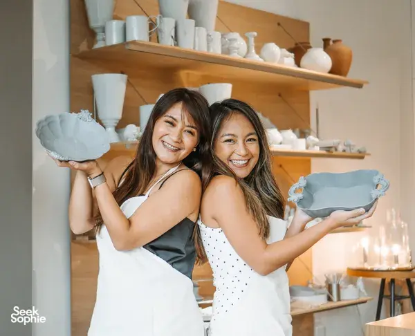Two smiling friends in aprons holding blue ceramic dishes they made in a Singapore pottery studio, antique-inspired European style