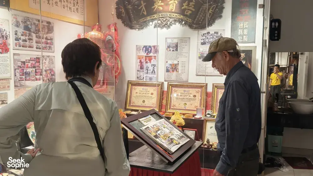 Visitors reading framed photos and certificates about lion dance and kung fu heritage in a Chinatown exhibition room, Ho Chi Minh City