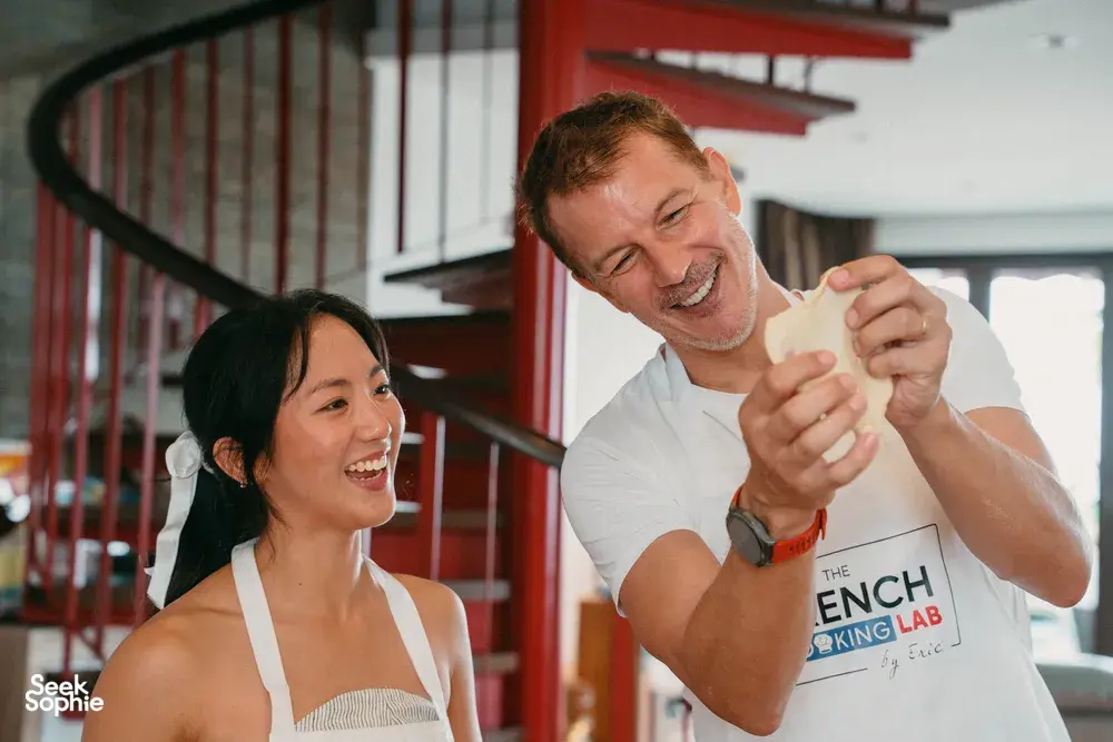 French chef showing croissant technique with a smiling participant in a Singapore cooking studio, both holding dough pieces