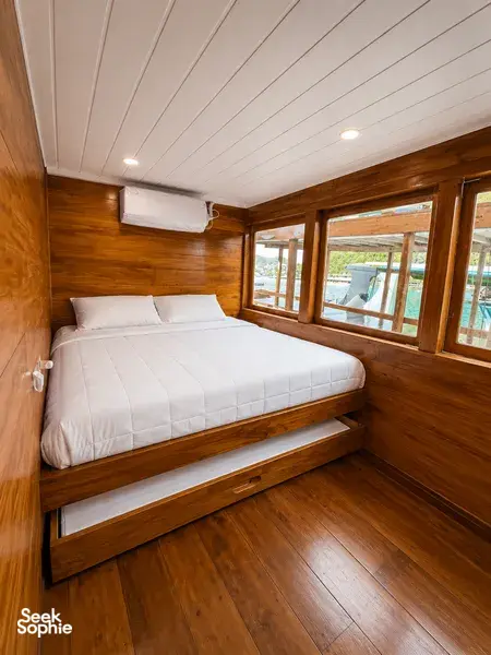 Wooden phinisi cabin with a neatly made double bed and large window views during a 3-day Komodo cruise in Labuan Bajo